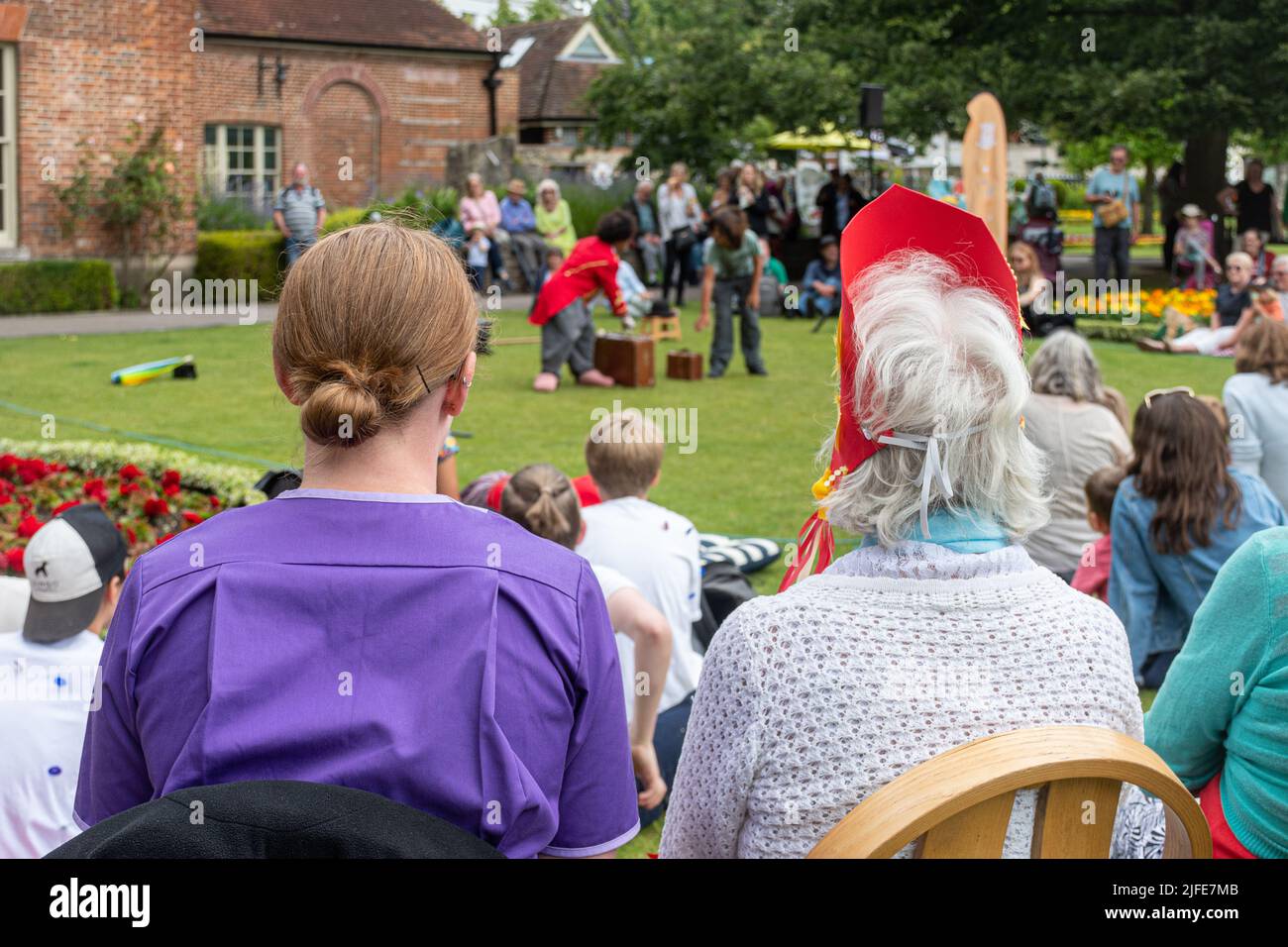Trabajador de cuidado sentado con una señora mayor en un parque, viendo a un animador de payasos, Inglaterra, Reino Unido. Cuidador que ayuda a personas mayores con actividades sociales. Foto de stock