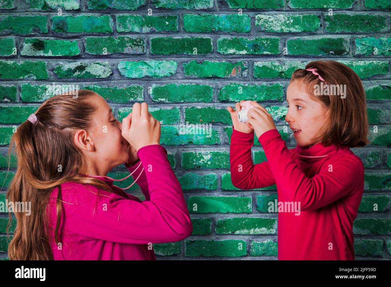 Una pareja de chicas pequeñas haciendo fotografías con la cámara de fotos de juguete, con una pared de ladrillo verde sobre el fondo Foto de stock