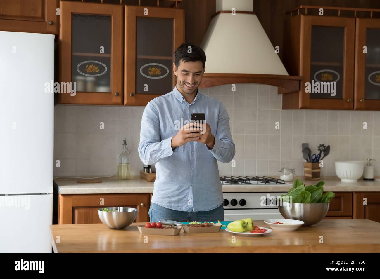 El hombre latino usa el teléfono celular mientras cocina el plato vegetariano en la cocina Foto de stock
