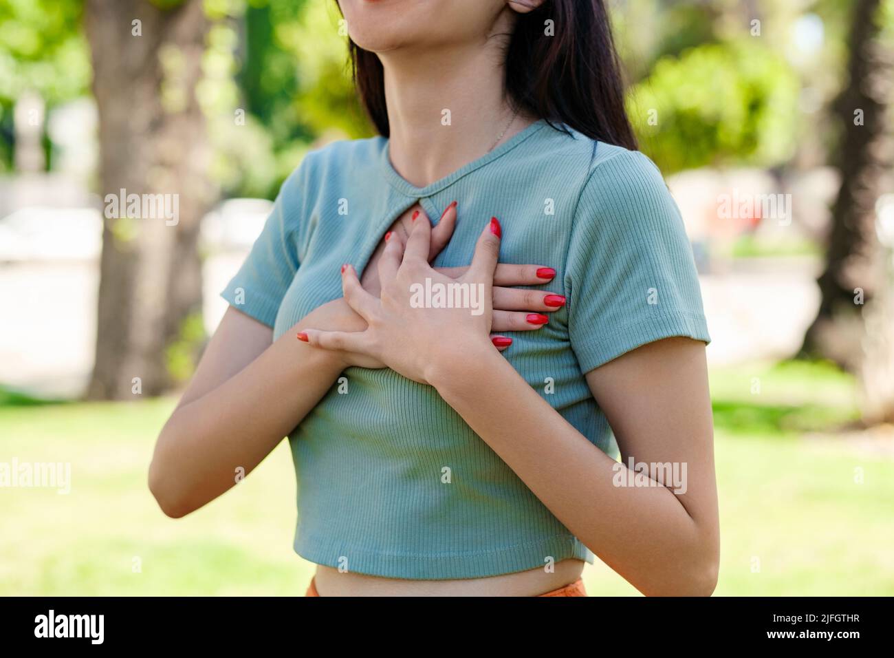 Joven mujer hermosa con camiseta turquesa en el parque de la ciudad, al  aire libre presiona las manos sobre el pecho y la expresión facial dolor,  teniendo un ataque al corazón Fotografía