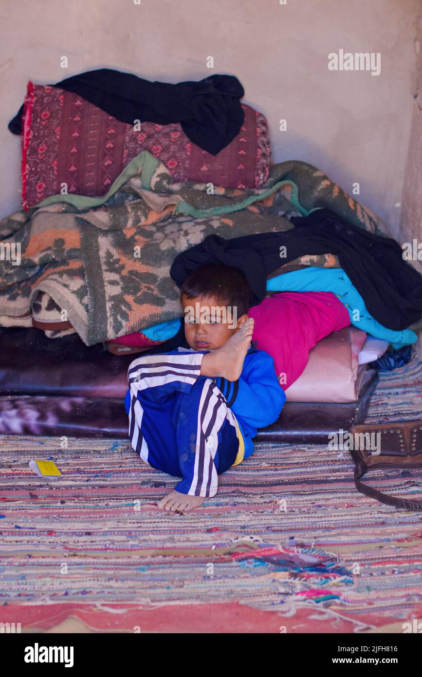 SHARM EL SHEIKH, EGIPTO - 31 2014 DE OCTUBRE: El pequeño niño egipcio se sienta sobre una alfombra poniendo la pierna sobre la rodilla apoyada en una cama desordenada con mantas y ropa. Niño pequeño Foto de stock