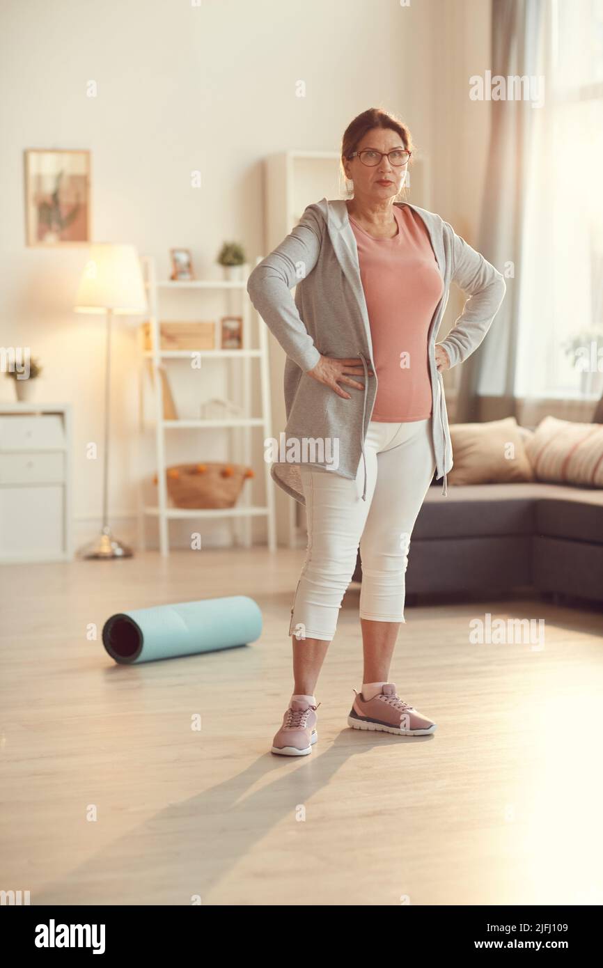 Retrato de una mujer mayor seria en deportiva y gafas de pie en el salón y con las manos en las caderas Fotografía de stock Alamy