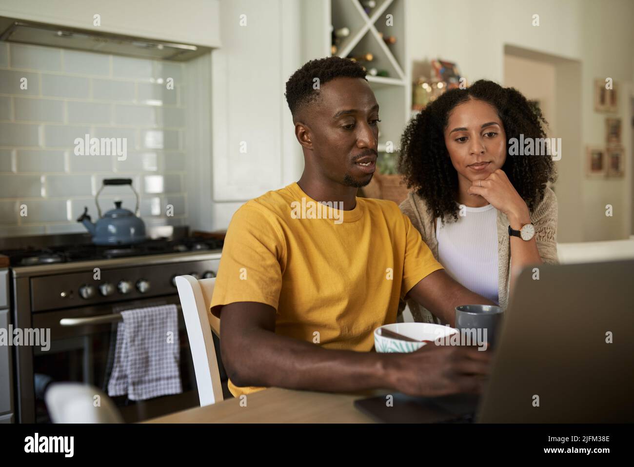 Una joven pareja multiétnica con un ordenador portátil durante el desayuno en una mesa de la cocina Foto de stock