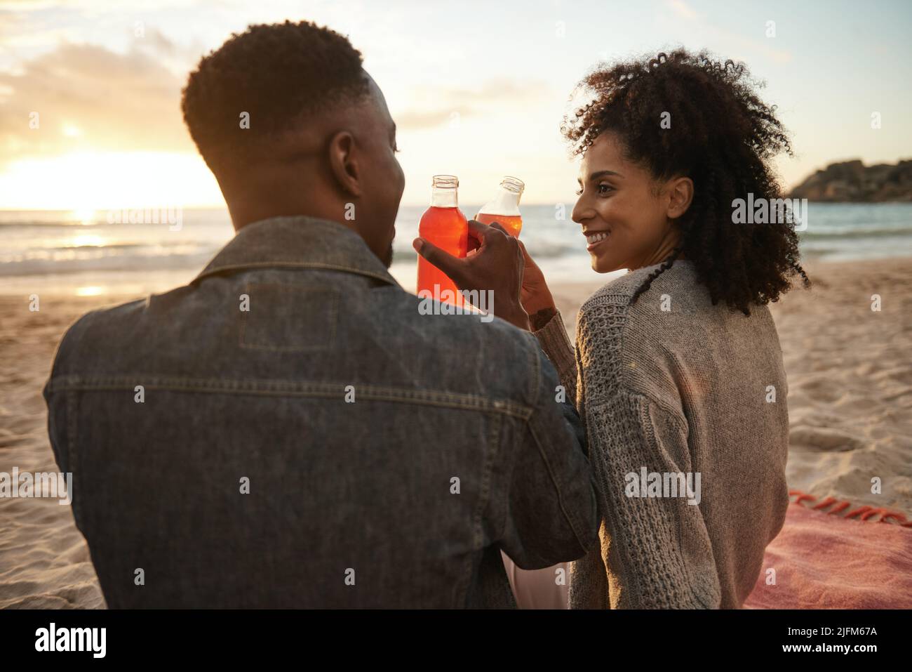 Sonriendo joven pareja multiétnica bebiendo jugo en una playa de arena al atardecer Foto de stock