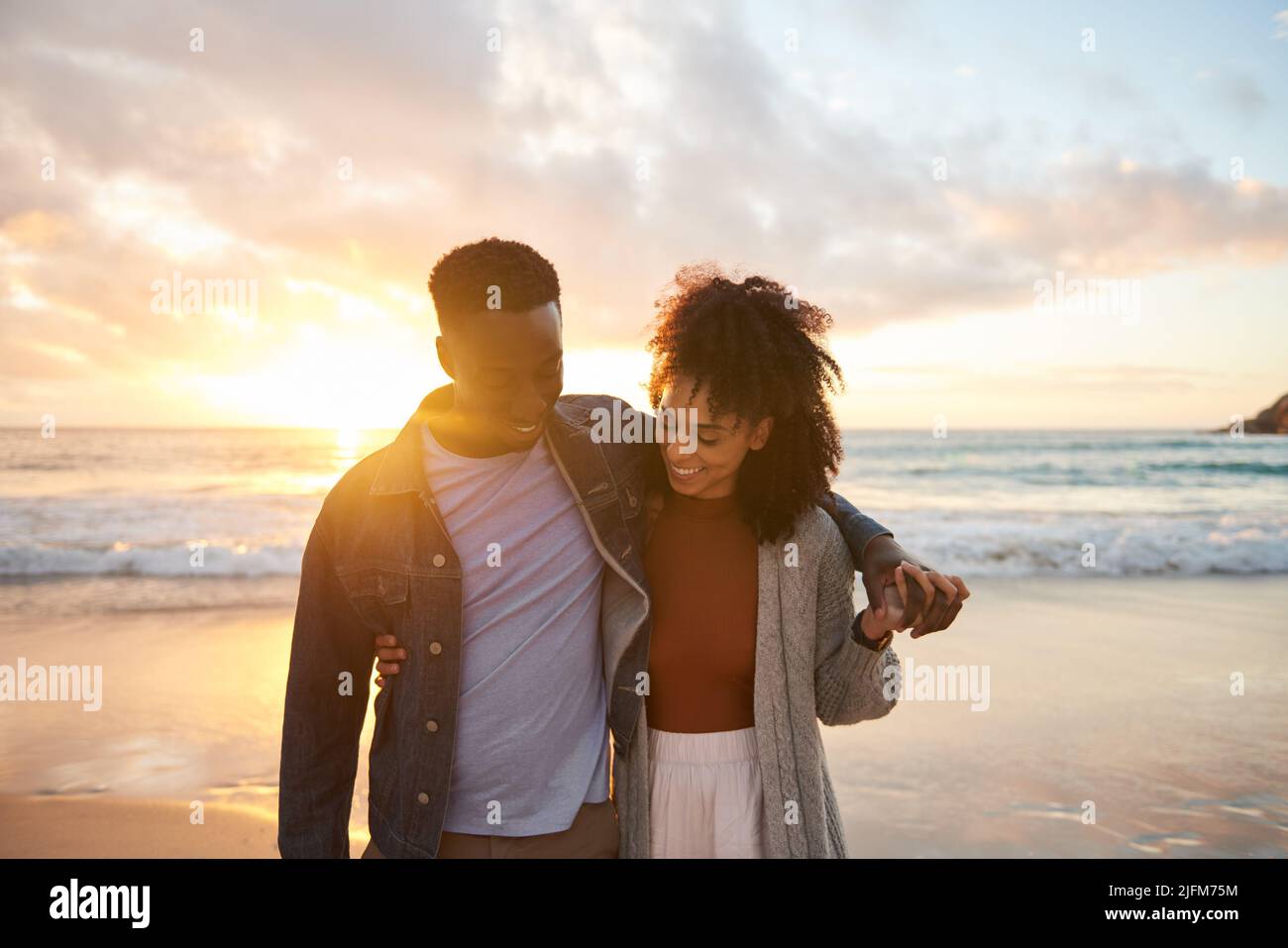 Sonriendo pareja multiétnica caminando de brazo en brazo en una playa al atardecer Foto de stock