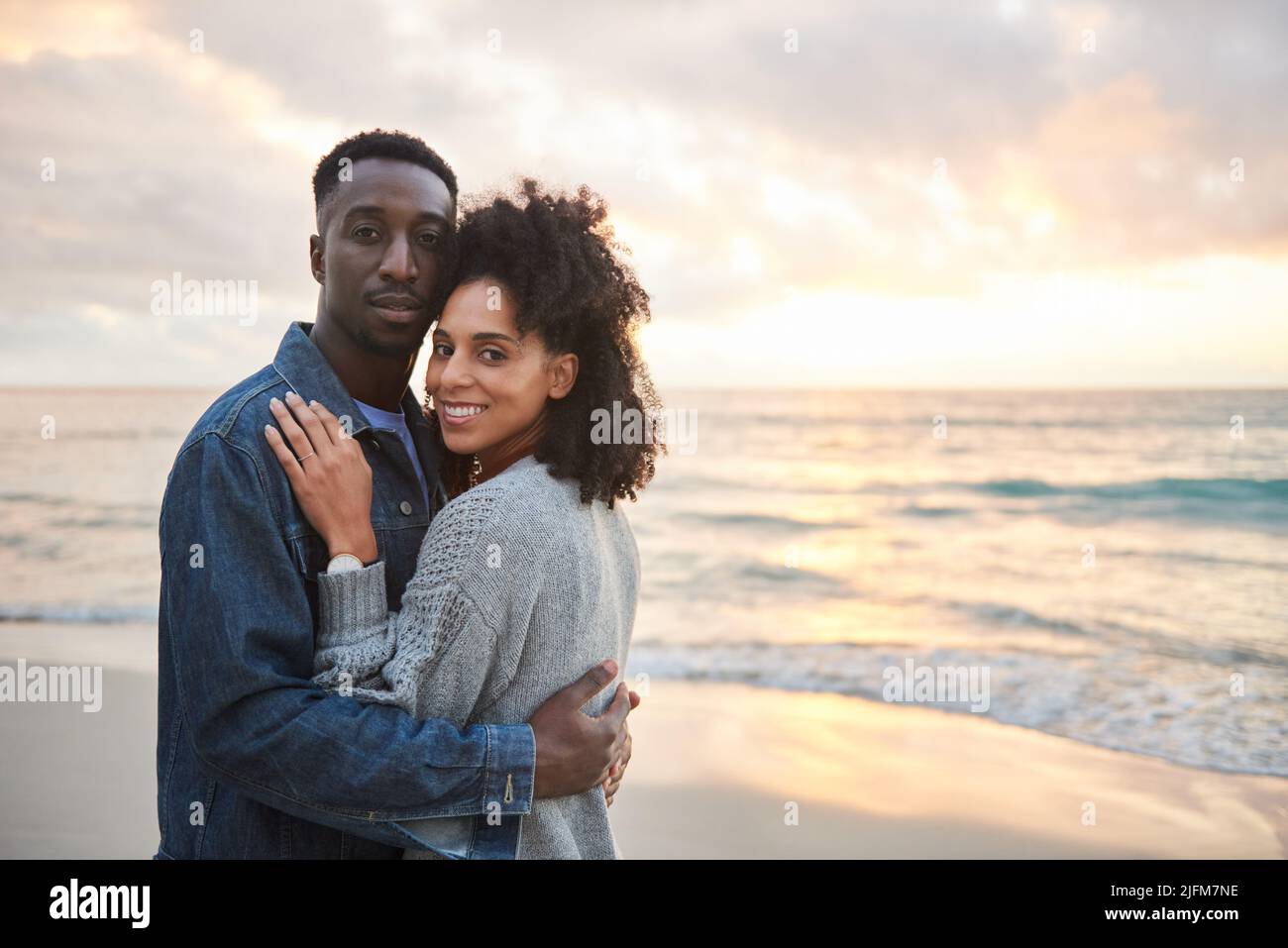 Joven pareja multiétnica de pie de brazo en brazo en una playa al atardecer Foto de stock