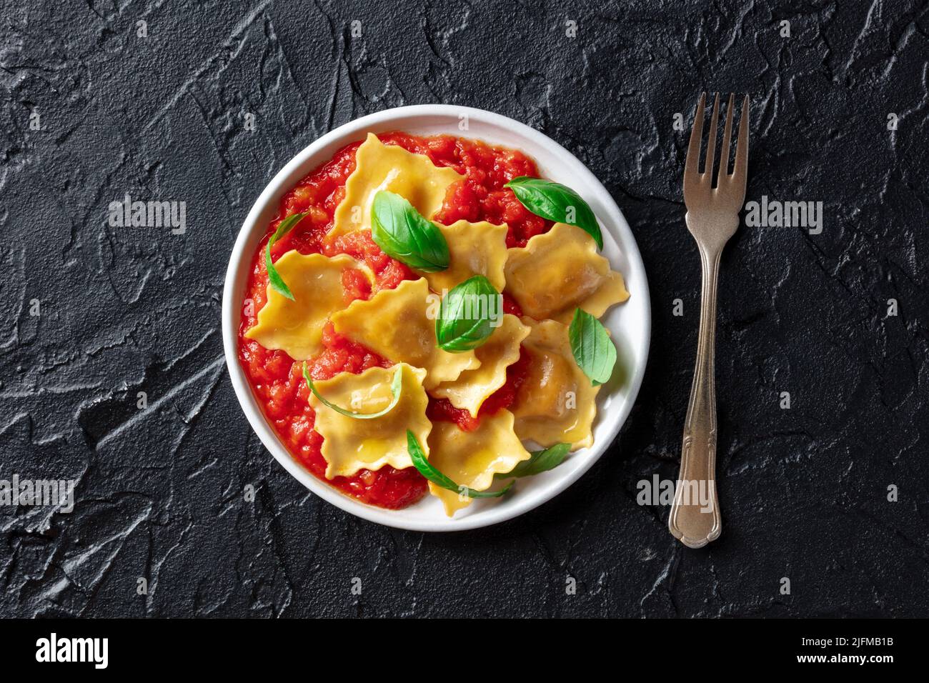 Ravioli con salsa de tomate y hojas frescas de albahaca en un plato, sobre la cabeza plana tiro sobre un fondo negro pizarra, con un tenedor Foto de stock