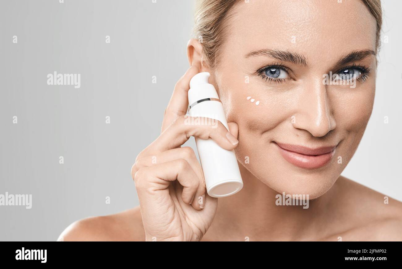 Mujer caucásica positiva aplica crema hidratante en su cara, sobre fondo gris. Crema facial hidratante piel, cuidado de la piel Foto de stock