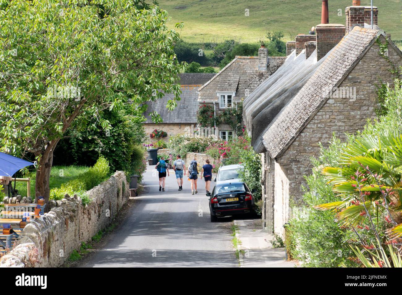 Kimmeridge, Dorset, Reino Unido. Cuatro jóvenes caminantes recorren la carretera principal en un pequeño pueblo inglés tradicional al comienzo de una caminata o paseo. Foto de stock