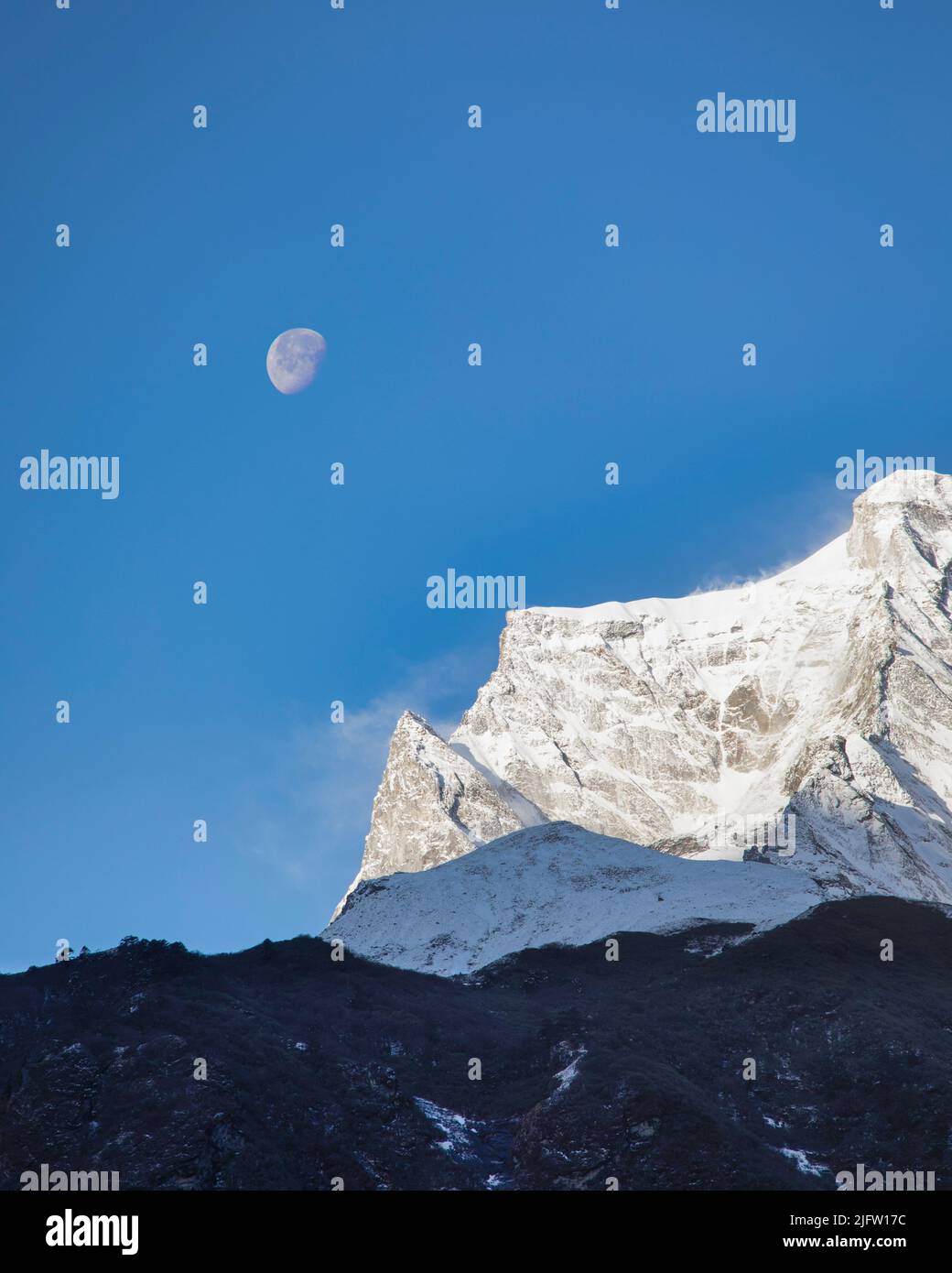 La luna permanece en la mañana mientras los vientos soplan nieve desde las elevaciones más altas de Kangtega en el Parque Nacional Sagarmatha, Nepal. Foto de stock