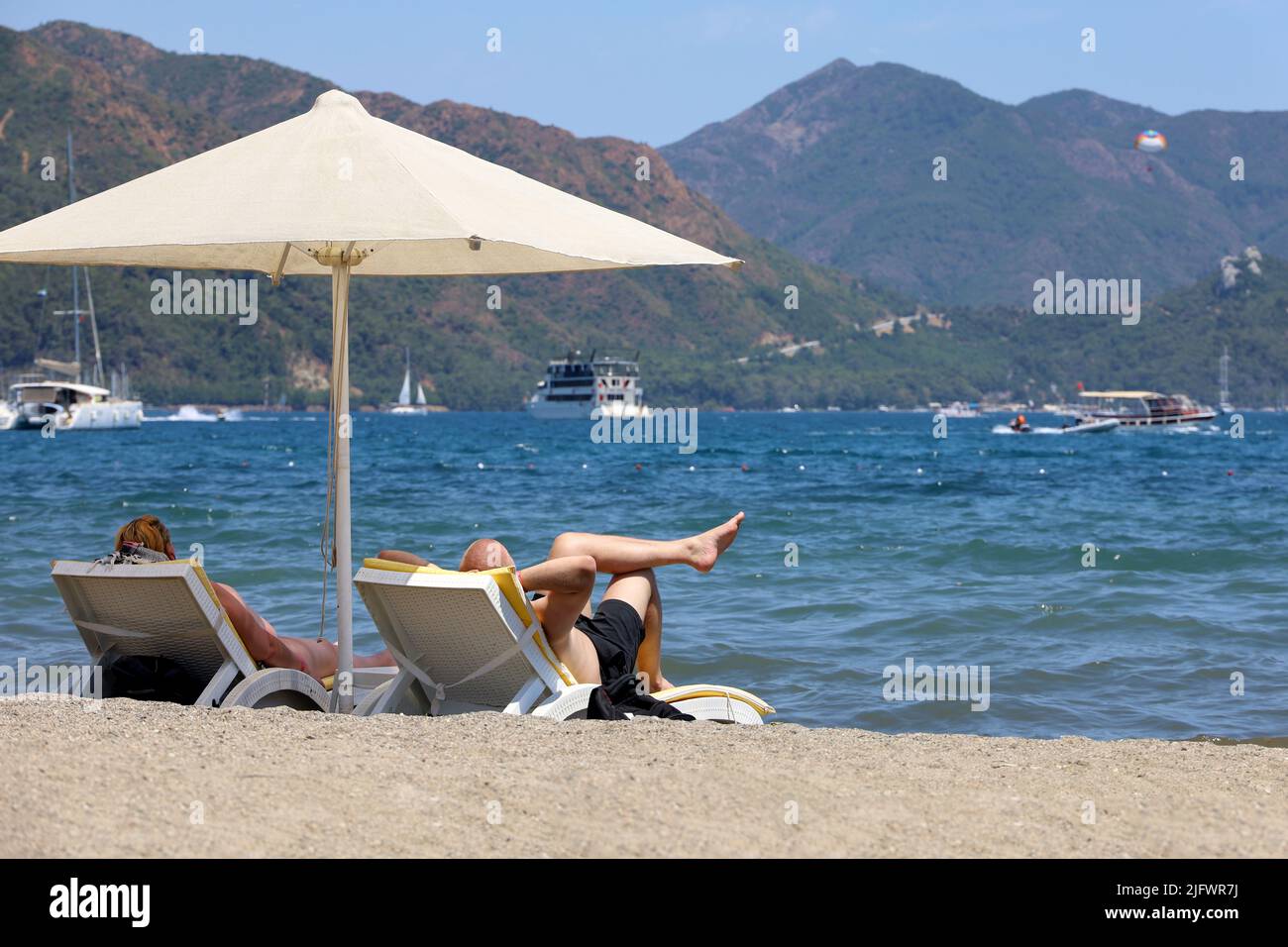 Pareja bronceándose en una playa en un resort de verano. Hombre y mujer tumbados en sillas de terraza bajo una sombrilla sobre las olas del mar y el fondo de la costa de montaña Foto de stock