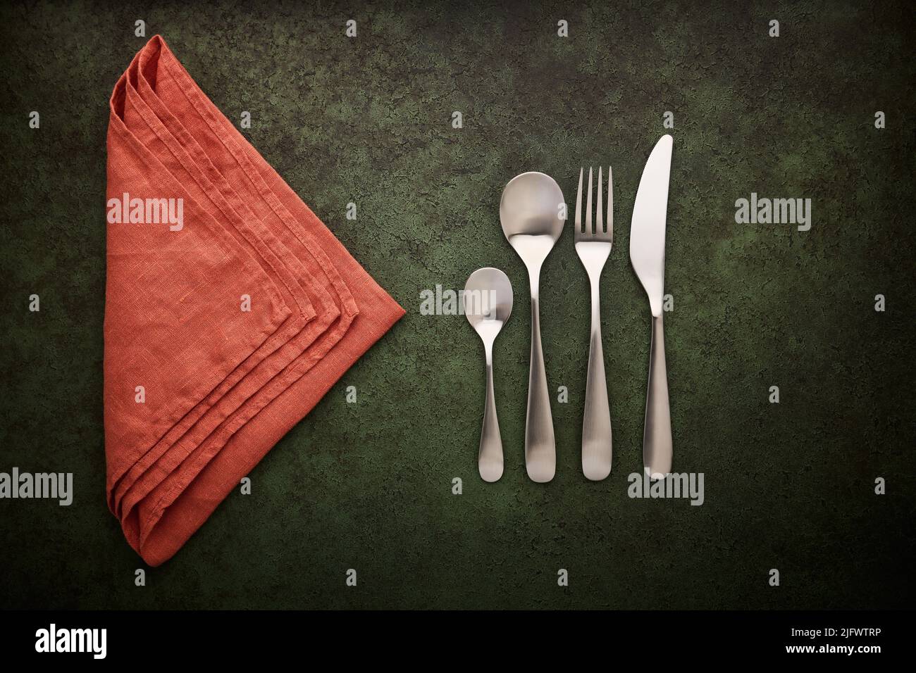 Un juego de cubertería y servilleta de lino de terracota sobre un fondo verde oscuro. Cucharas, tenedor y cuchillo, puesta plana Foto de stock