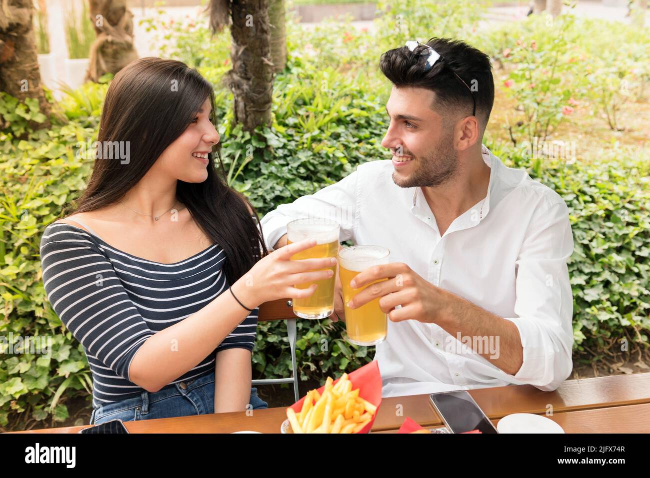 Pareja joven romántica que se topan con cerveza fría al aire libre en un parque o jardín sonriendo y feliz mientras celebran su amor Foto de stock