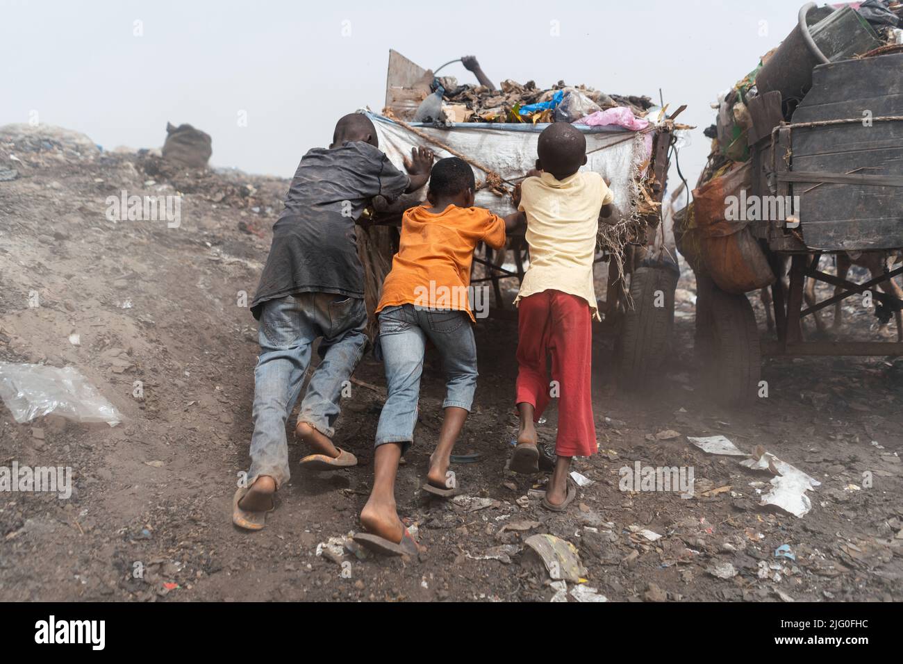 Tres muchachos negros de la calle empujando un carro pesado de basura encima de un montón en un vertedero urbano; gestión informal de residuos en África Foto de stock
