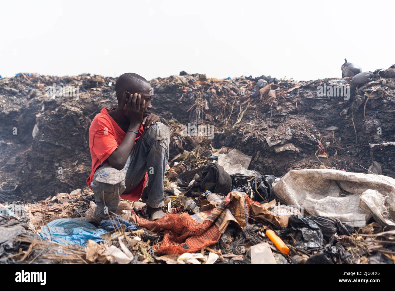 Pobre chico de los barrios marginales africanos sentado frente a una gran pila de basura en medio de un enorme vertedero urbano después de un día de trabajo en busca de reciclable Foto de stock