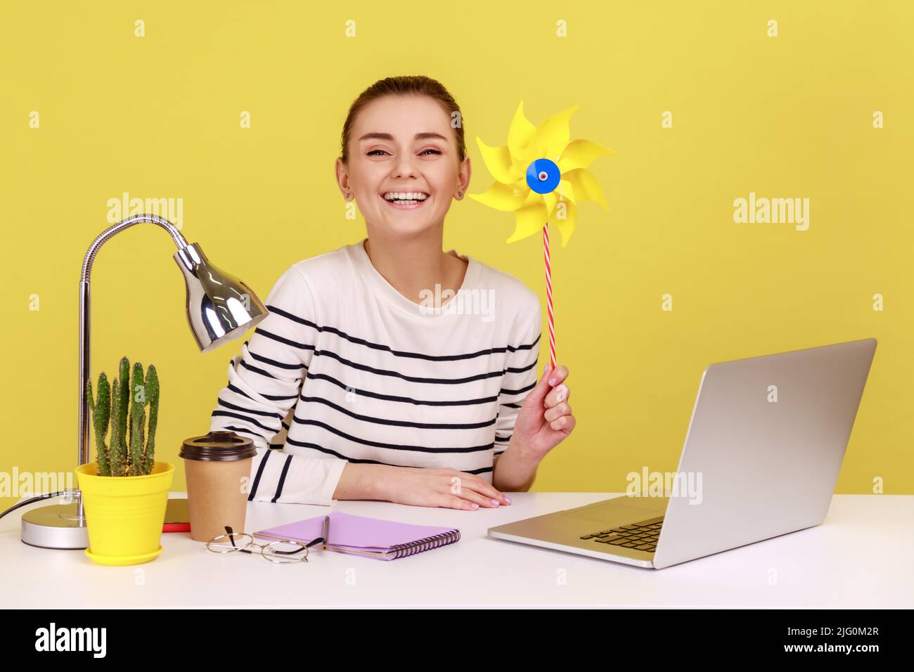 Origami molino de mano. Retrato de feliz sonrisa despreocupada mujer sosteniendo molino de viento de papel, juguete de rueda de alfiler en palo, divertirse en el lugar de trabajo. Estudio de interior grabado aislado sobre fondo amarillo. Foto de stock