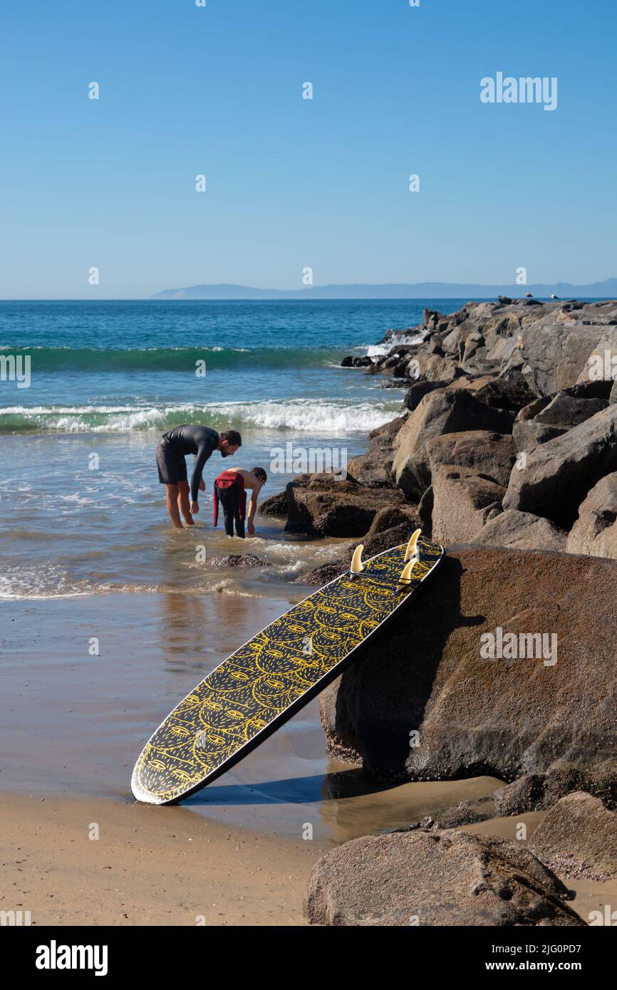 Tabla de surf contra la barrera de roca construida para proteger o retener la playa con el hombre y el niño en el mar n el fondo Newport Beach Southern California Foto de stock