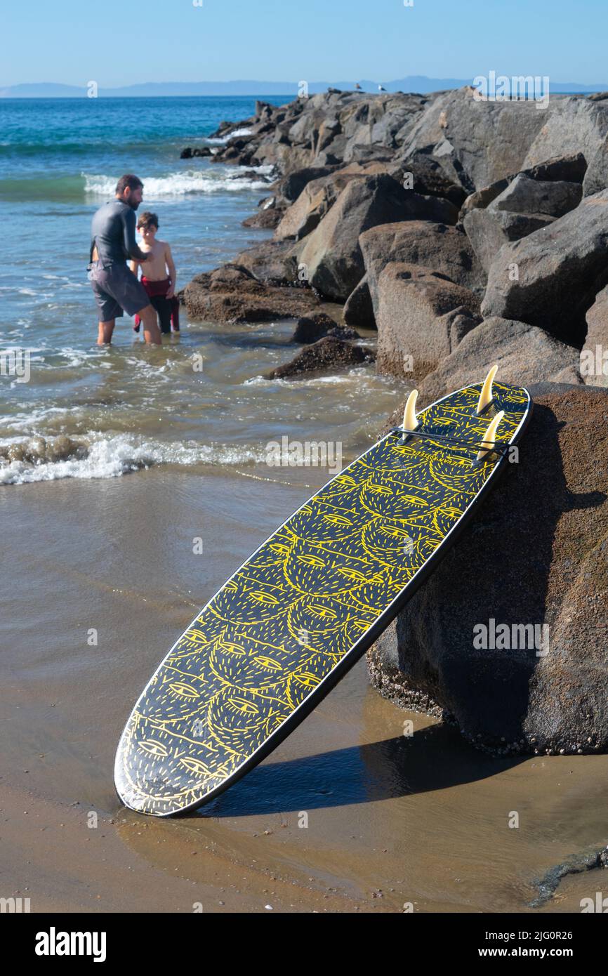 Tabla de surf contra la barrera de roca construida para proteger y retener la playa con el hombre y el niño en el mar en el fondo Newport Beach Southern California Foto de stock