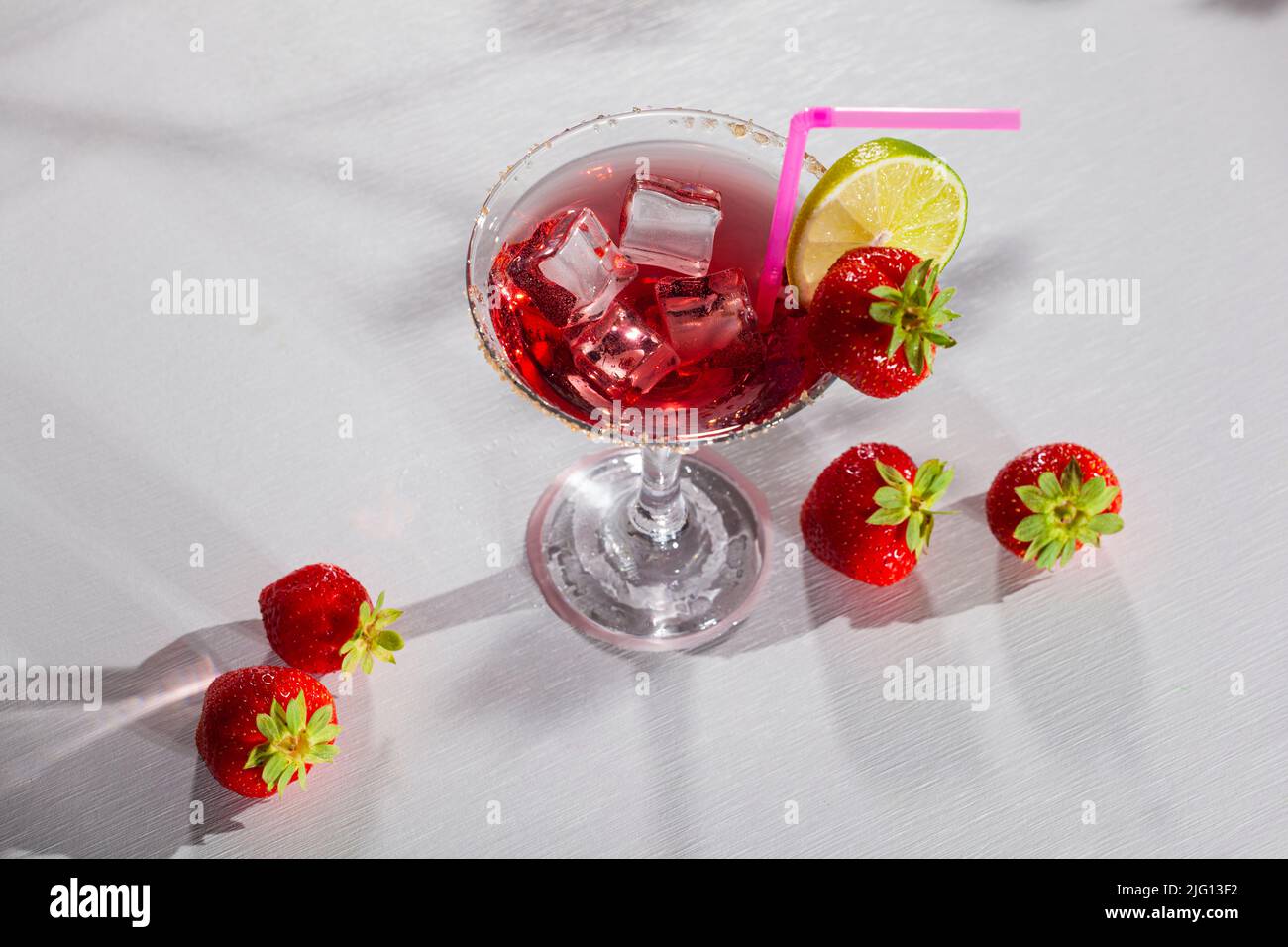 Delicioso cóctel. Bebida roja dulce y fría en un elegante vaso con cubitos de hielo, fresa y lima sobre mesa blanca Foto de stock