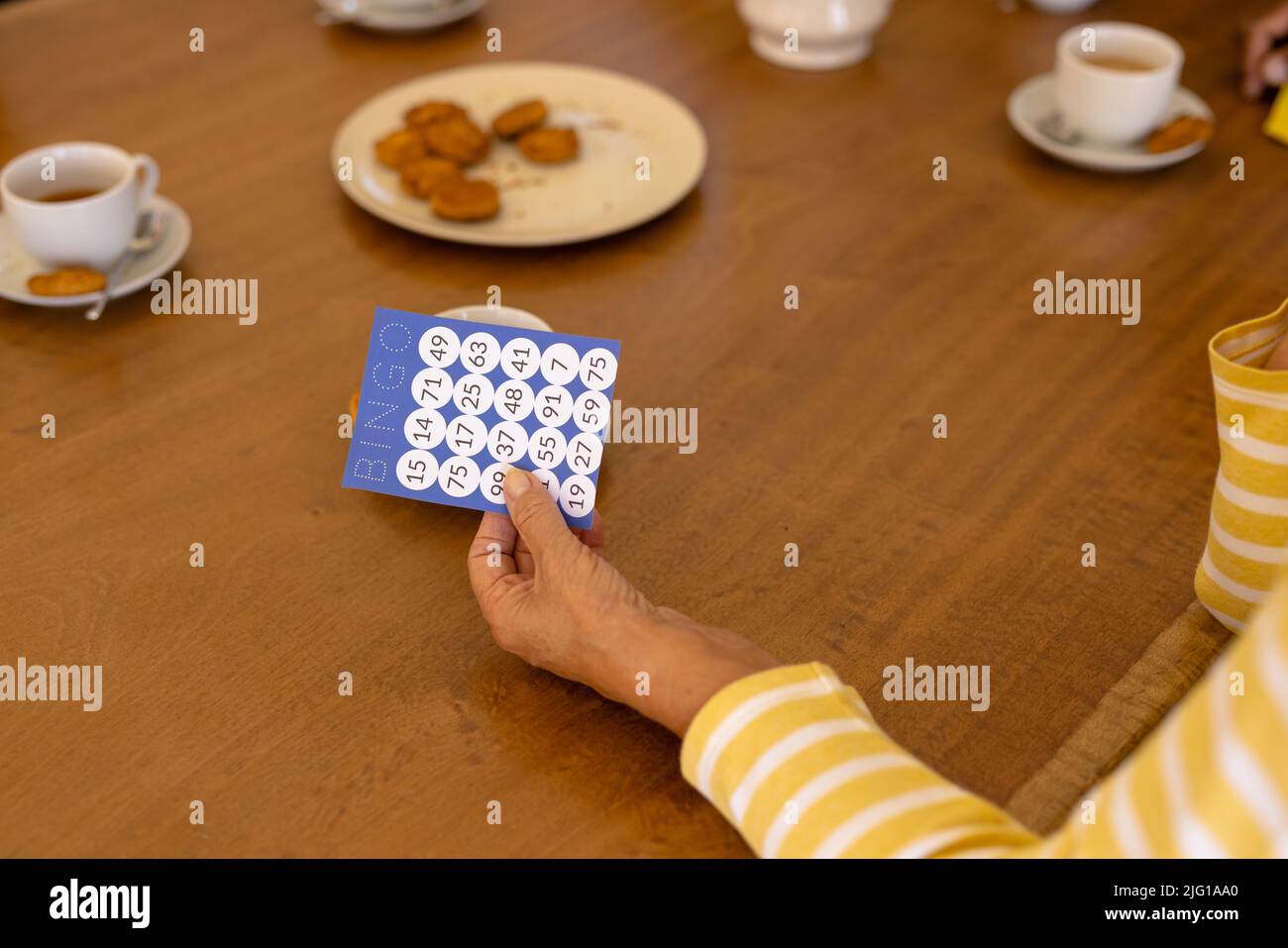 Mano recortada de una mujer mayor biracial sosteniendo una tarjeta de bingo en la mesa del comedor en un hogar de ancianos Foto de stock