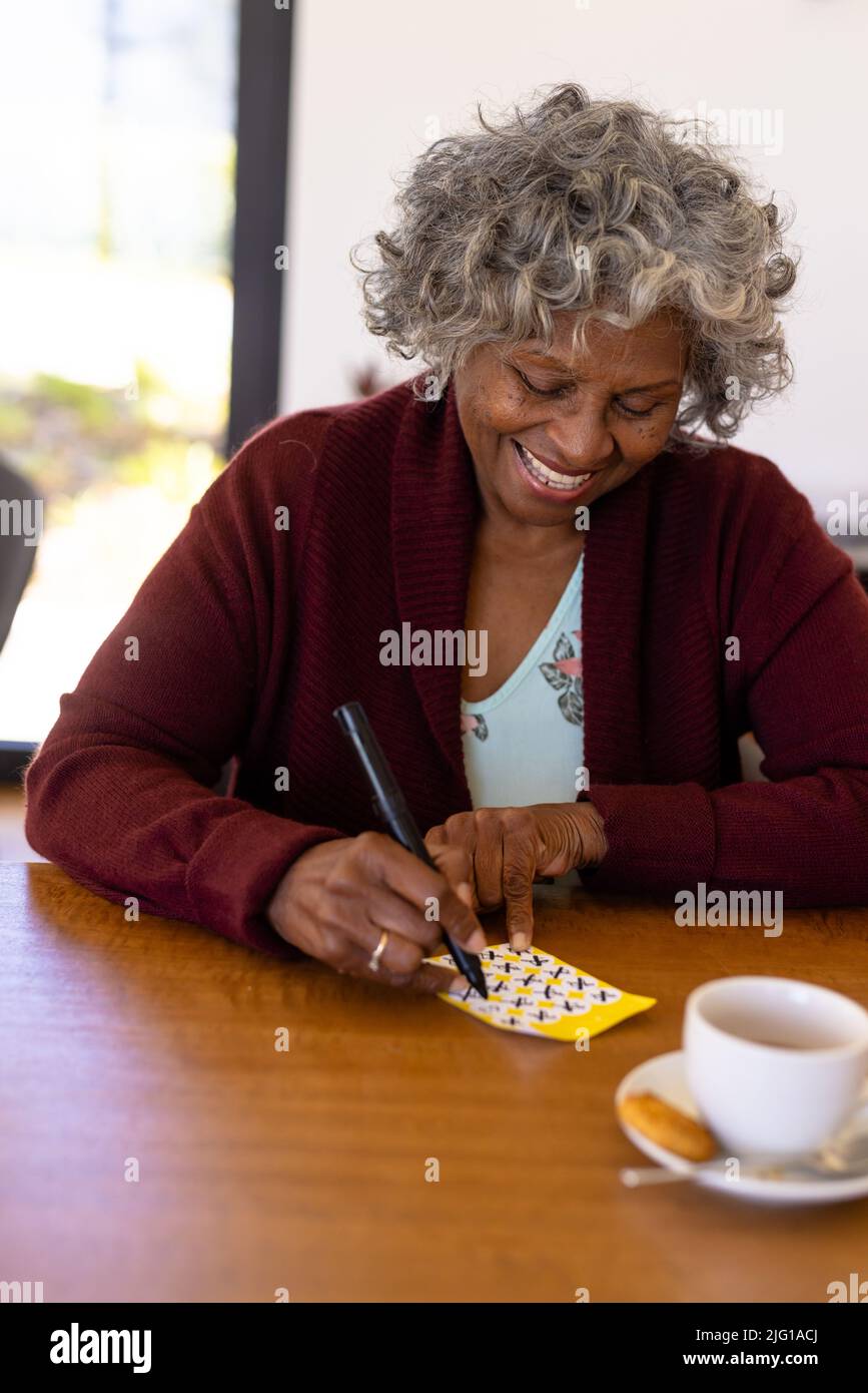 Sonriente mujer afroamericana que marca los números en la tarjeta de bingo sobre la mesa de comedor Foto de stock