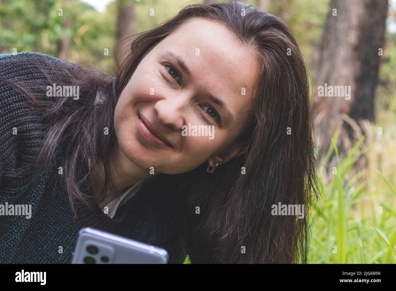 Retrato de una joven sonriente en un parque y mirando la cámara, sosteniendo el smartphone Foto de stock