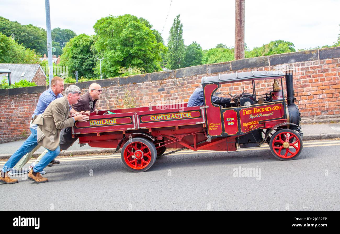 Tres hombres empujando un vagón de motor de vapor reducido sobre una colina en el pueblo de Cheshire, en Lymm, Inglaterra, después de haber perdido energía Foto de stock