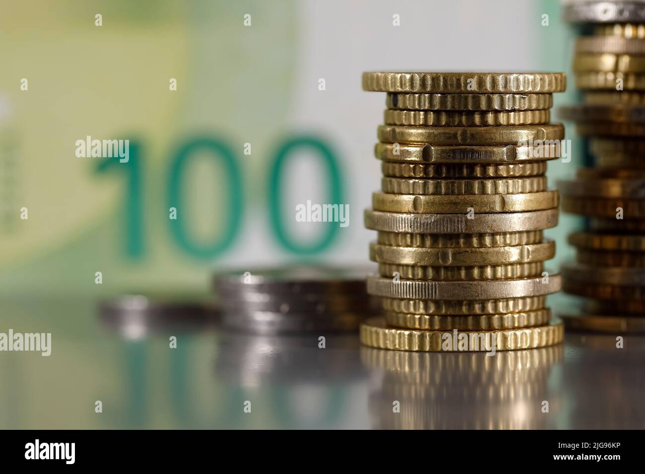 Se trata de monedas en euros que se han apilado sobre una superficie gris y plana y son visibles contra un billete borroso de 100 euros. Foto de stock