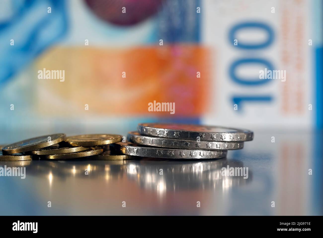 Esta pequeña pila de varias monedas se ha colocado sobre una superficie gris plana y es visible sobre el fondo de la nota borrosa de 100 francos suizos. Foto de stock