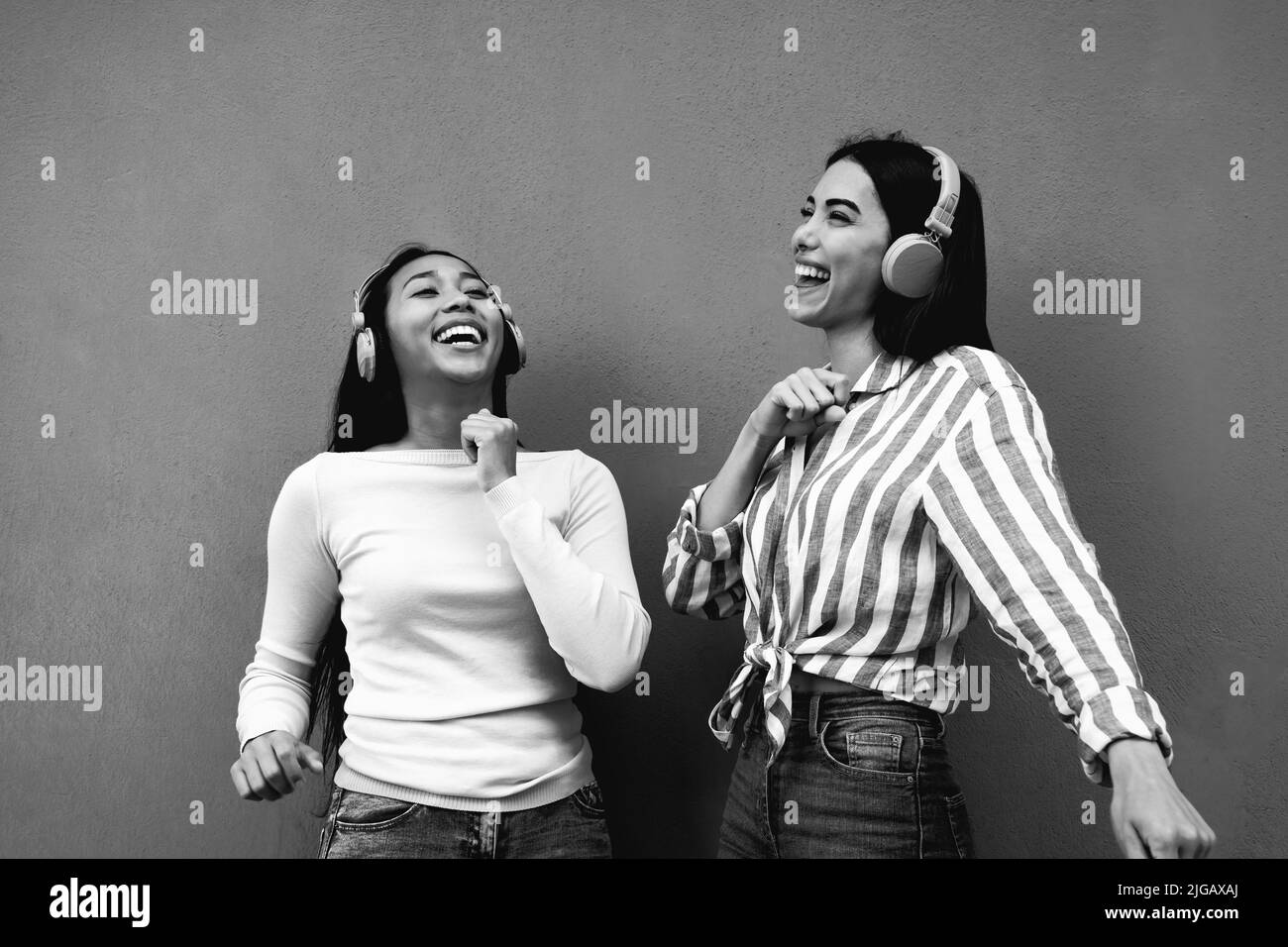 Jóvenes chicas latinas felices divirtiéndose bailando y escuchando música con auriculares inalámbricos al aire libre - Edición en blanco y negro Foto de stock