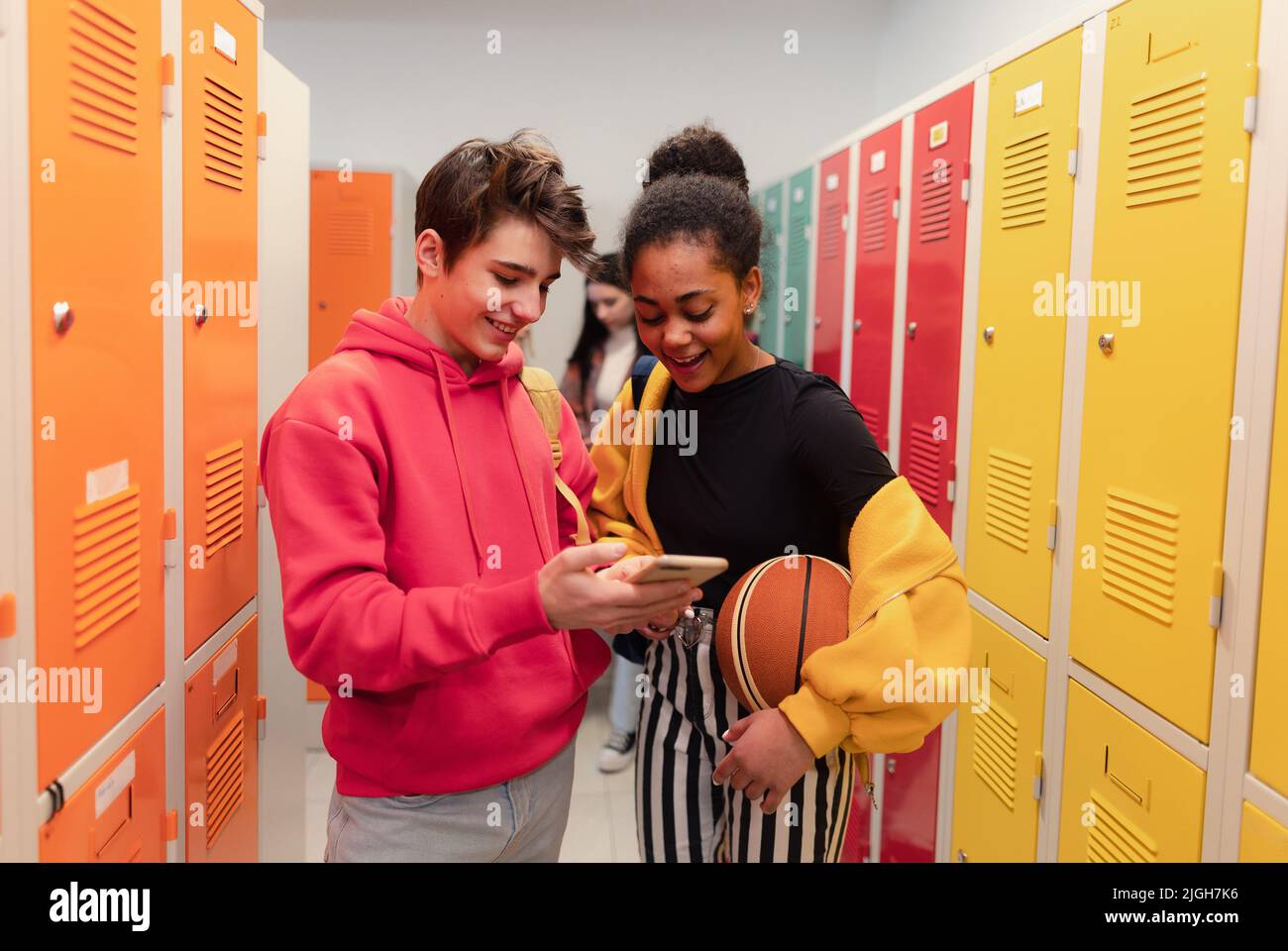Jóvenes estudiantes de secundaria de pie cerca de un armario en el pasillo del campus hablando y utilizando el teléfono inteligente. Foto de stock