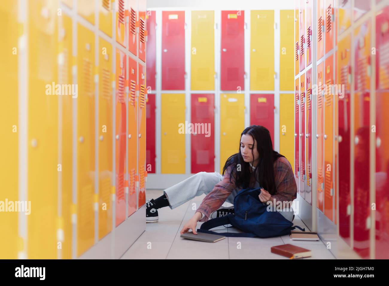 Estudiante adolescente sentado en el pasillo cerca de casilleros coloridos y empacando libro a mochila en el pasillo del campus, concepto de regreso a la escuela. Foto de stock