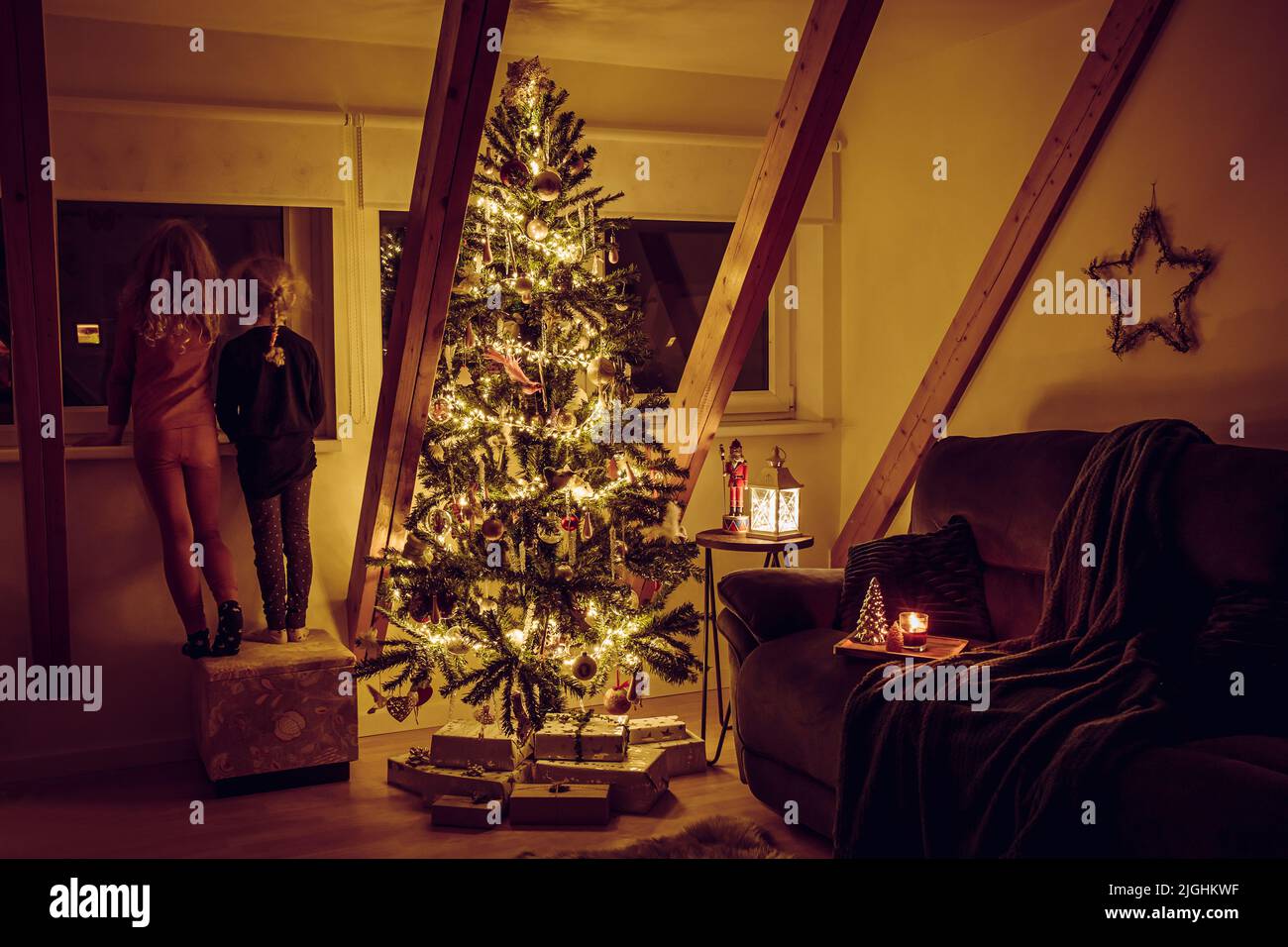 Hogar sala de estar con árbol de Navidad decorado, luces y decoraciones colgando, regalos bajo el árbol y dos hermanas mirando por la ventana. Foto de stock