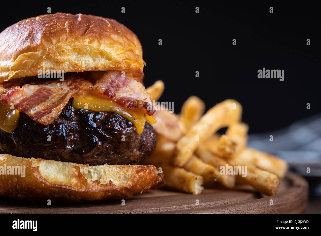Primer plano de hamburguesa con queso de tocino sobre pan tostado con patatas fritas en una bandeja de madera. Fondo negro con espacio de copia Foto de stock