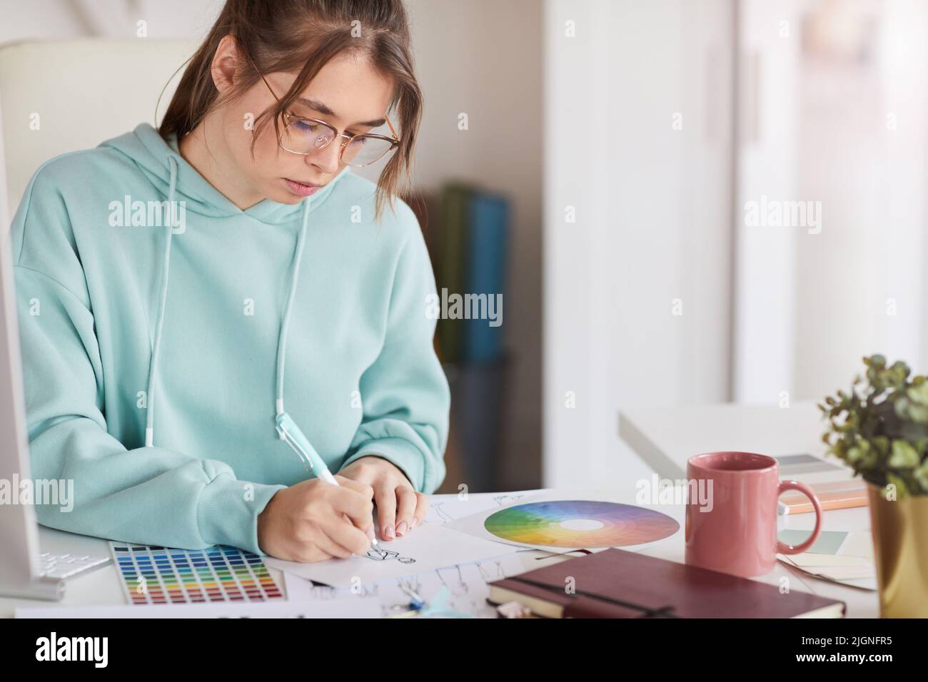 Serio concentrado joven diseñadora en la sudadera con capucha azul verdoso sentado en la mesa y dibujo de la moda bosquejo con pluma Foto de stock