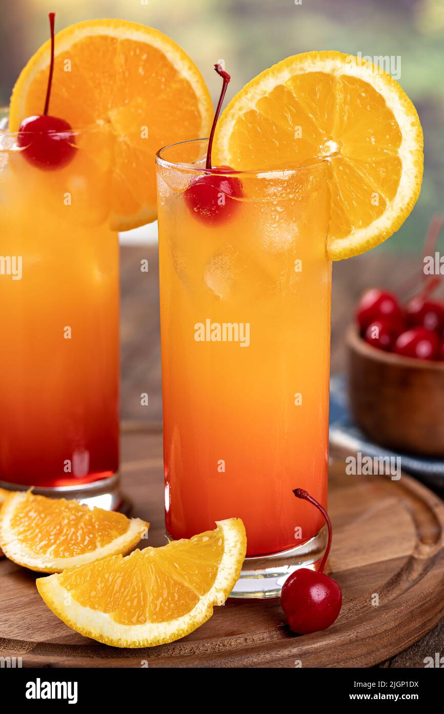 Dos cócteles Tequila Sunrise con rodaja de naranja y cereza en una bandeja rústica de madera Foto de stock