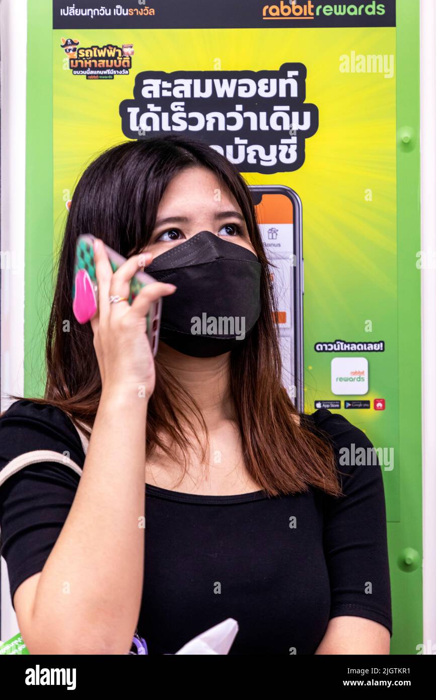 Pasajero tailandés con máscara de facemask utilizando el teléfono delante del anuncio en el Skytrain BTS, Bangkok, Tailandia Foto de stock