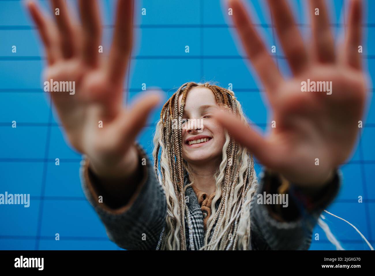 Una adolescente muy ligera bloqueando la cámara con las manos delante de un panel azul que cubre la pared. Ella está usando una chaqueta gris a cuadros. Foto de stock