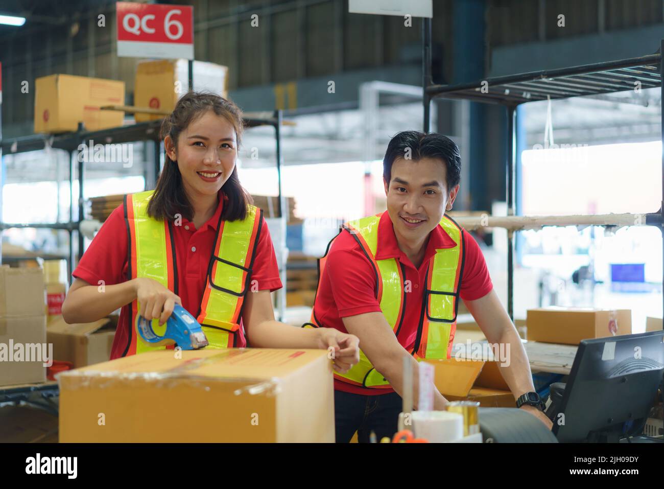 Trabajadora asiática embalando caja de cartón con dispensador de pistola de cinta en almacén. Empleado tailandés que empaqueta mercancías en almacén industrial grande. Foto de stock