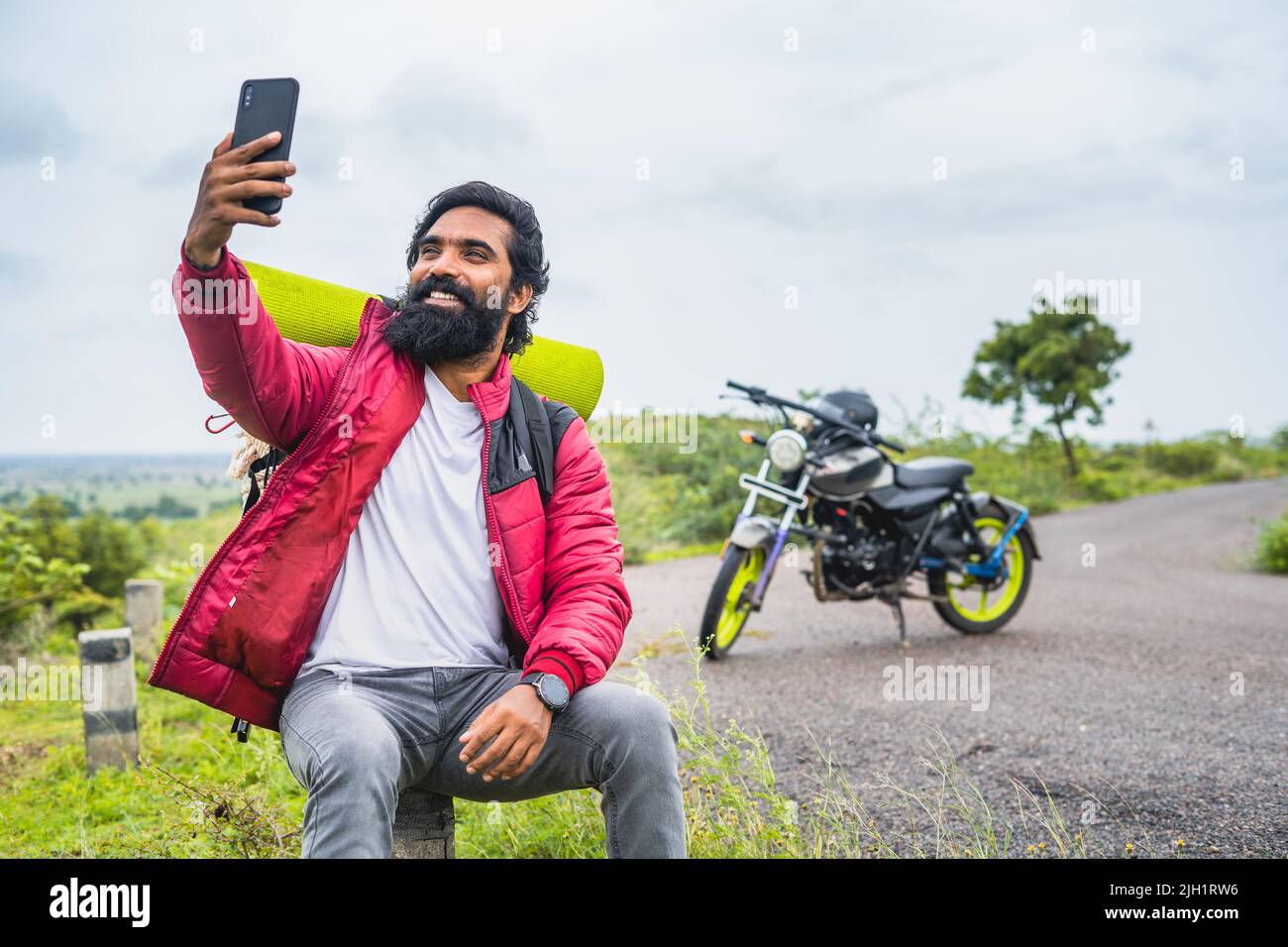 Un hombre de barba sonriente y feliz haciendo videollamadas en el teléfono móvil delante de la moto mientras se sienta en la carretera: Concepto de tecnología, redes sociales Foto de stock