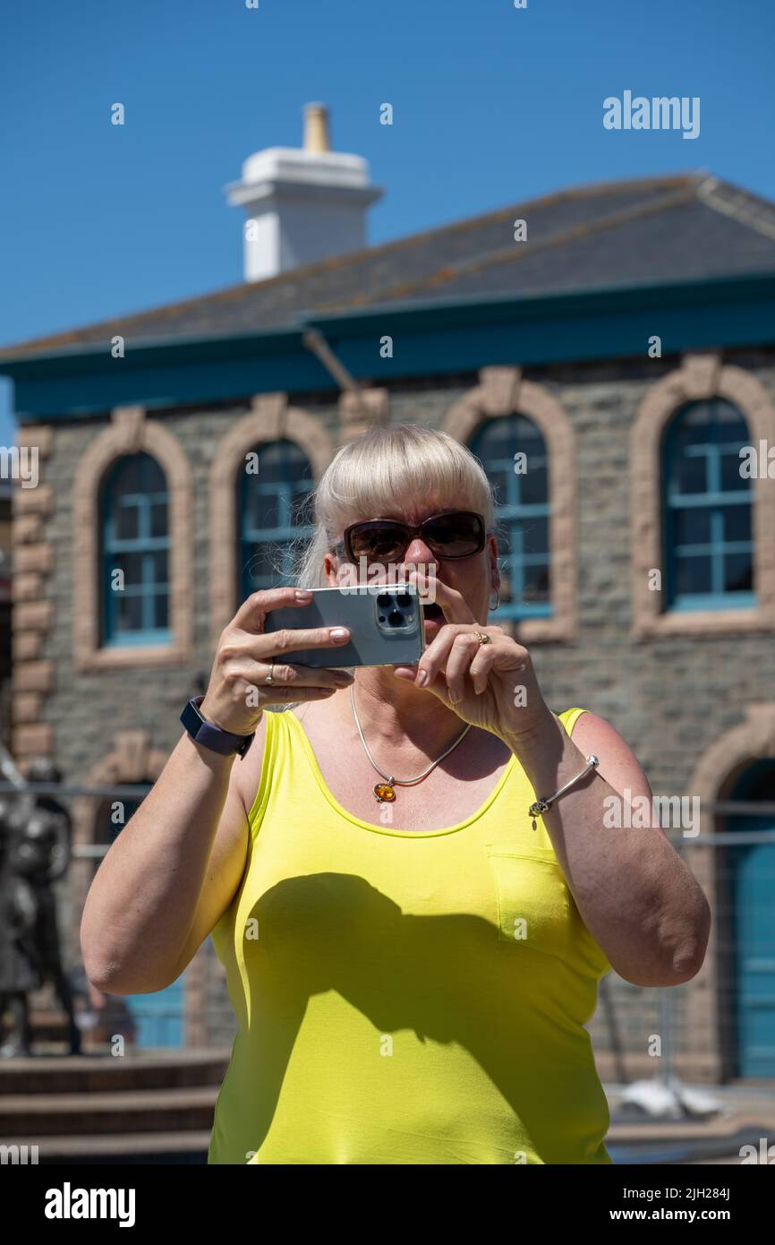 Turista adulta que lleva gafas de sol y una parte superior amarilla haciendo fotografías en un teléfono móvil mientras está de vacaciones en verano. Foto de stock
