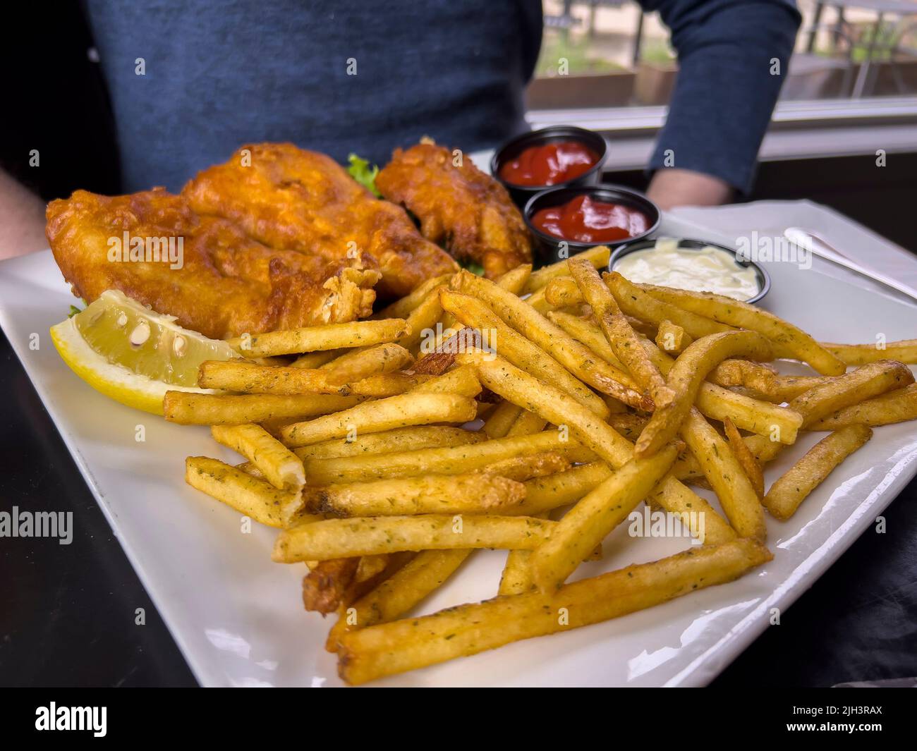 Vista de cerca de pescado crujiente y patatas fritas en una mesa en un restaurante frente a un hombre caucásico Foto de stock