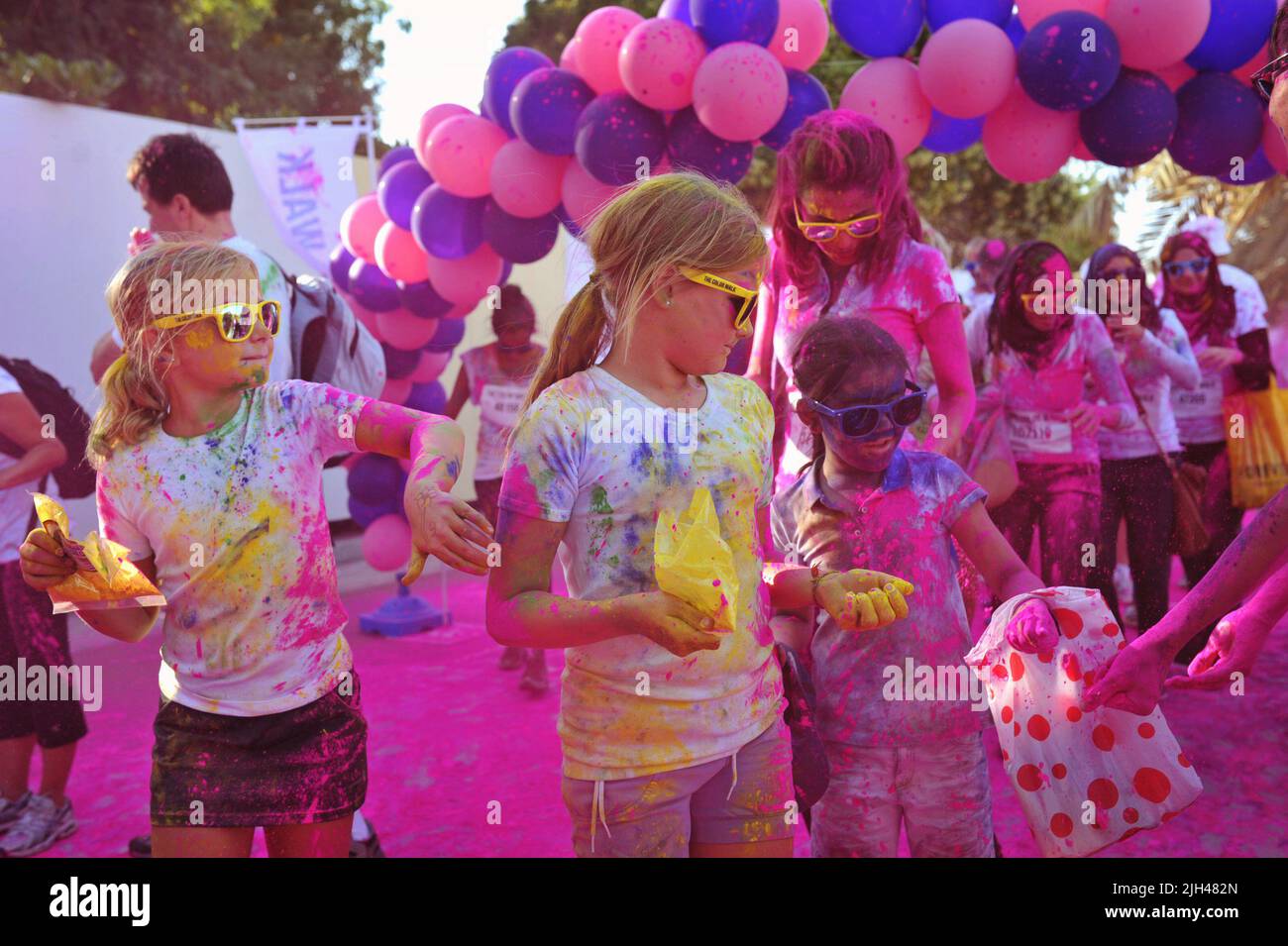 Chicas jóvenes rubias con tonos amarillos se detienen para agarrar polvo más coloreado junto a un arco de globo rosa en el Paseo del Color en Dubai, Emiratos Árabes Unidos. Foto de stock