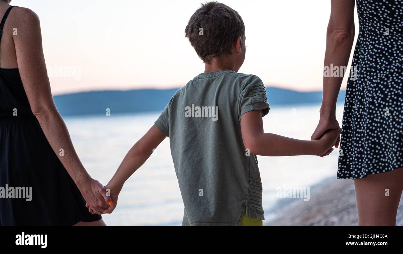 Vista desde atrás de dos madres, una pareja lesbiana, sosteniendo a su hijo por sus manos caminando juntos por la playa. Foto de stock