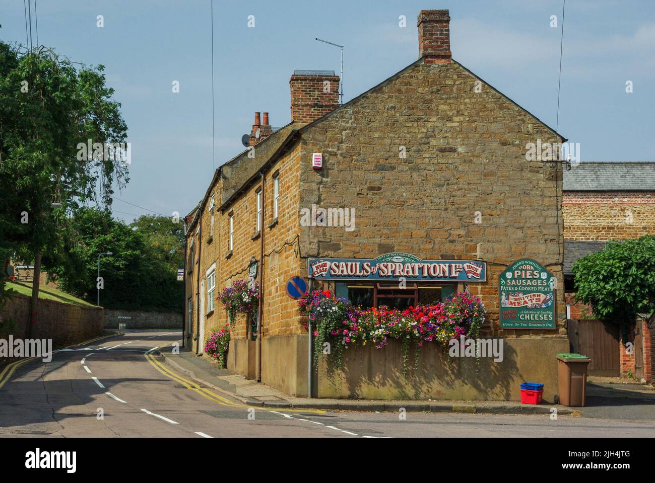 Escena callejera en verano en el bonito pueblo de Spratton, Northamptonshire, Reino Unido; edificios de piedra de color miel incluyendo la carnicería de Sauls. Foto de stock