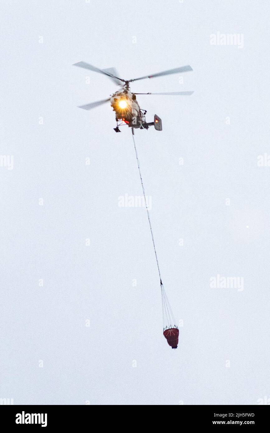 Un helicóptero contra incendios lleva un contenedor de agua para extinguir un incendio en un edificio de producción Foto de stock