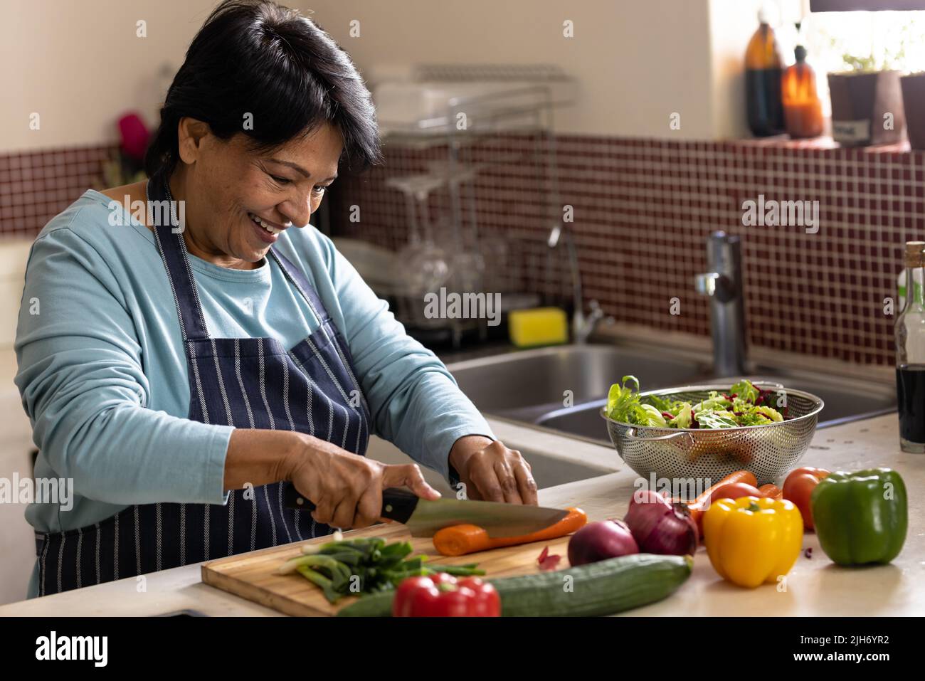 Sonriendo mujer madura biracial con pelo corto usando delantal cortar verduras a bordo en la cocina Foto de stock