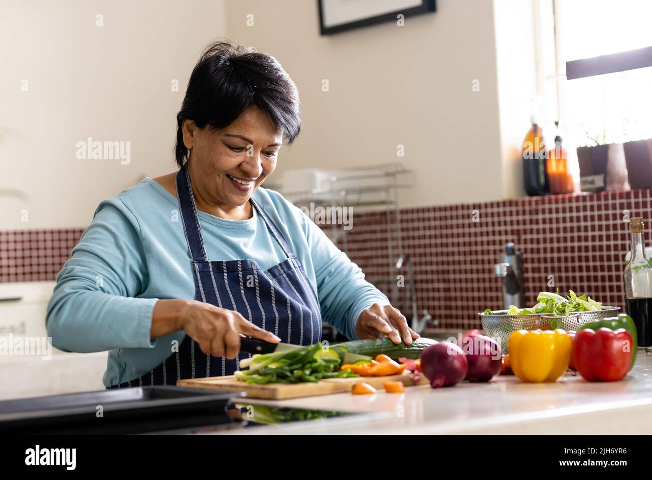 Sonriente mujer madura biracial con pelo corto usando delantal que corta verduras a bordo en la cocina Foto de stock