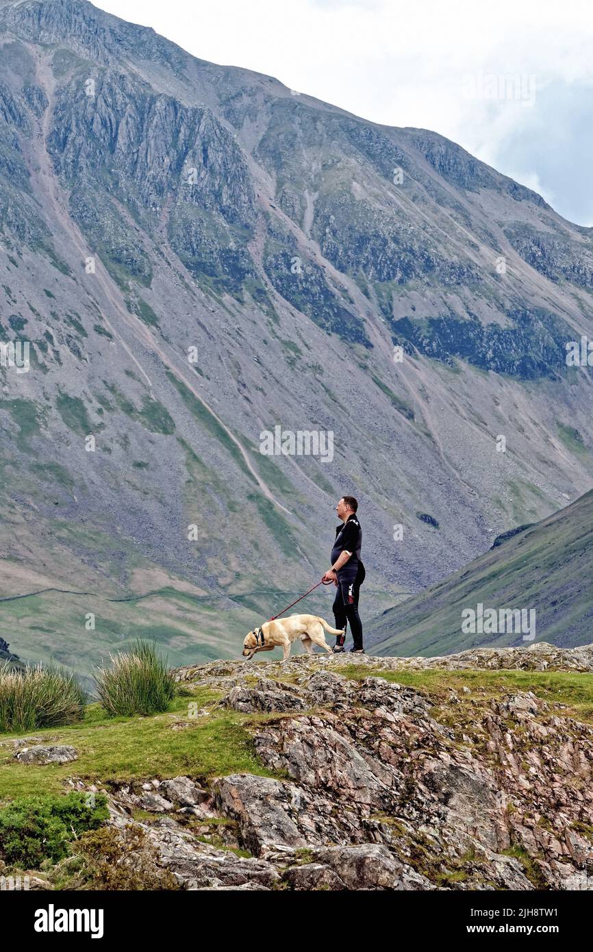 Un hombre de mediana edad con perro parado en un afloramiento rocoso con la montaña Great Gable en el fondo admirando la vista en Wasdale Cumbria, Inglaterra, Reino Unido Foto de stock
