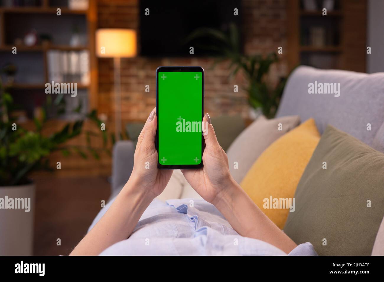 Vista sobre el hombro de una mujer joven sosteniendo un smartphone con pantalla verde de efecto doble en modo horizontal.Mujer tumbada en el sofá en casa por la noche, viendo Foto de stock
